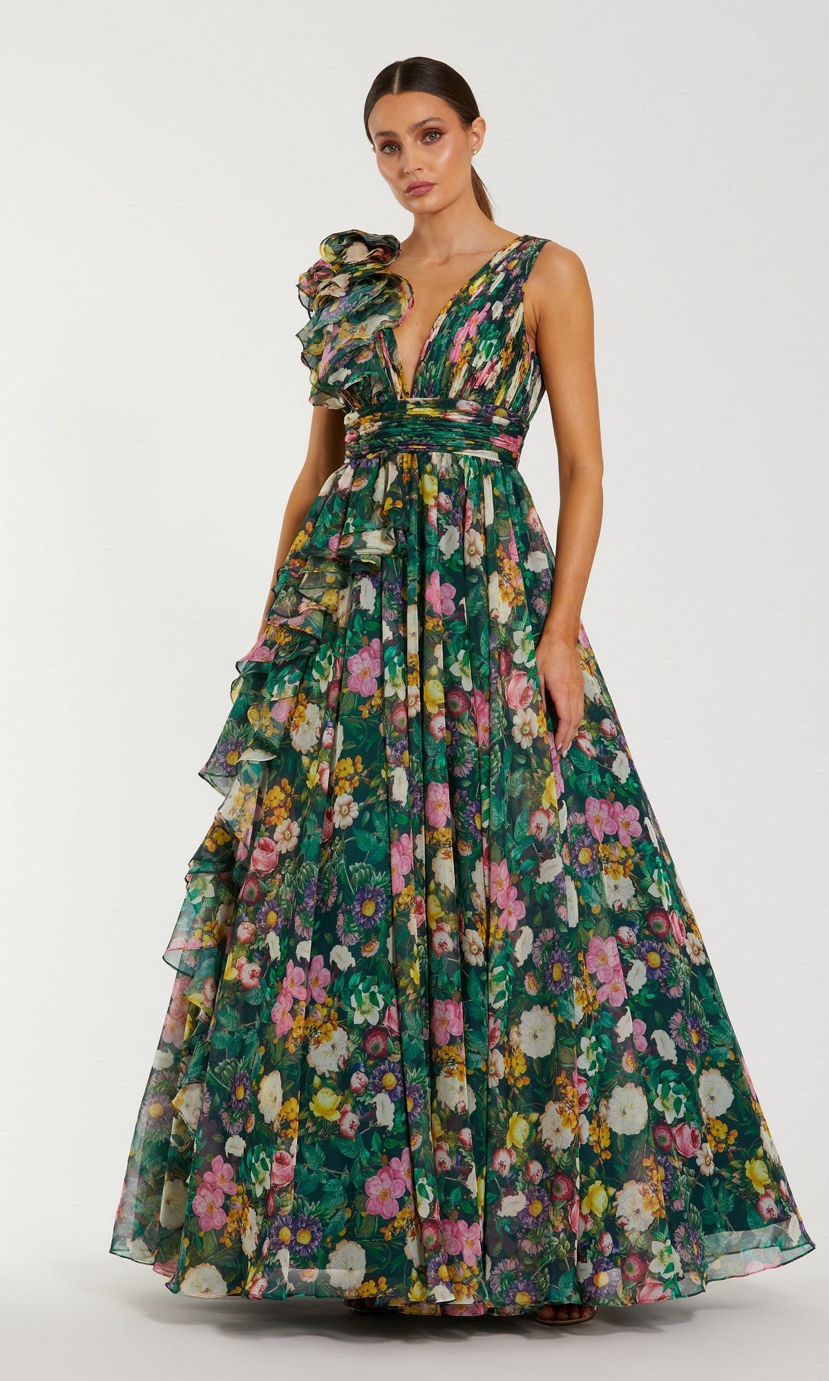 A woman wears the Mac Duggal 68947 Floral-Print Long Formal Dress in Deep Green Multi, with sleeveless floral-print chiffon, ruffled details, and a deep V neckline, standing against a plain light background.